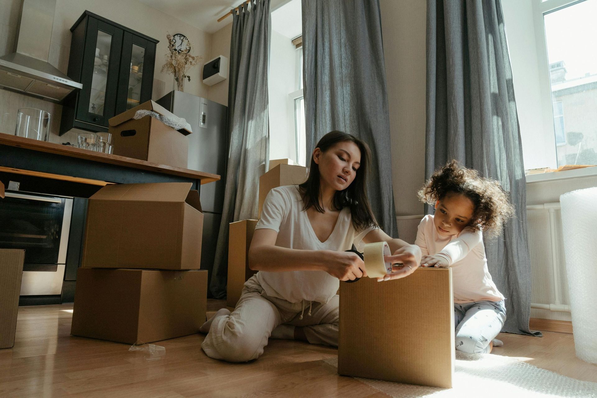 A person and a child packing cardboard boxes together in a sunlit living room with grey curtains.