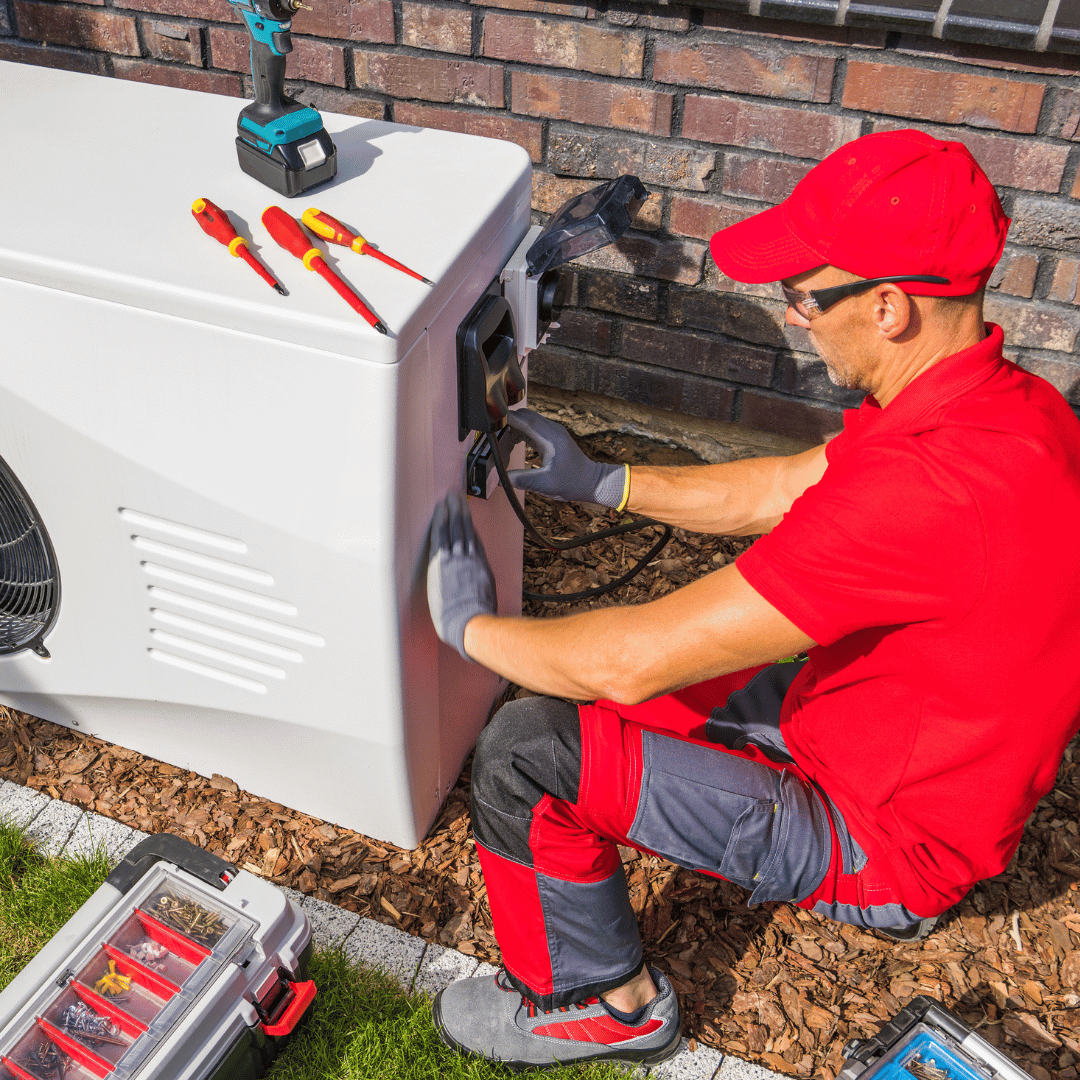 A man in a red shirt is sitting on the ground working on an air conditioner.