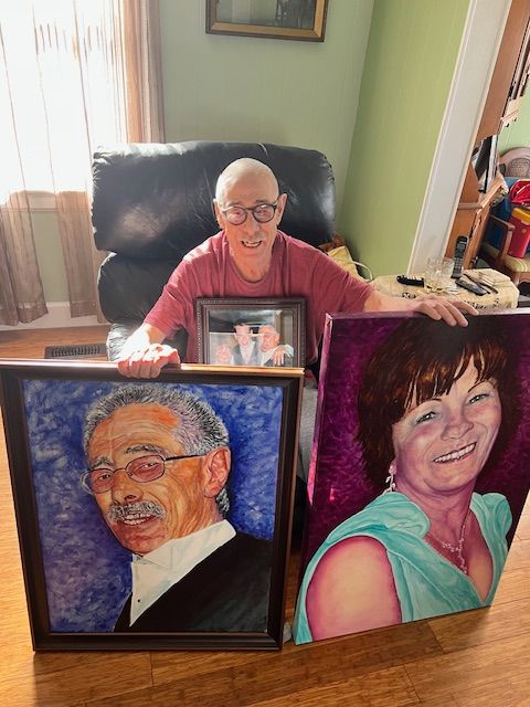 A man is sitting in a chair holding two paintings of a man and a woman.