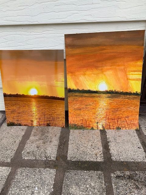 Two paintings of a sunset over a body of water are sitting on a tiled floor.