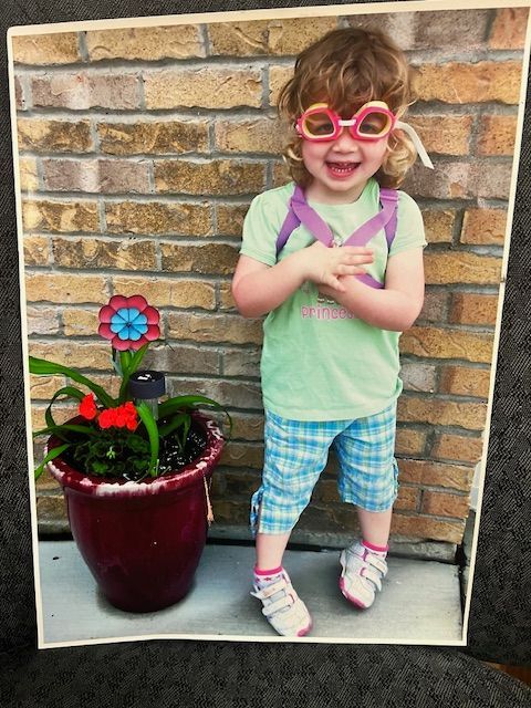 A little girl wearing sunglasses stands next to a potted plant