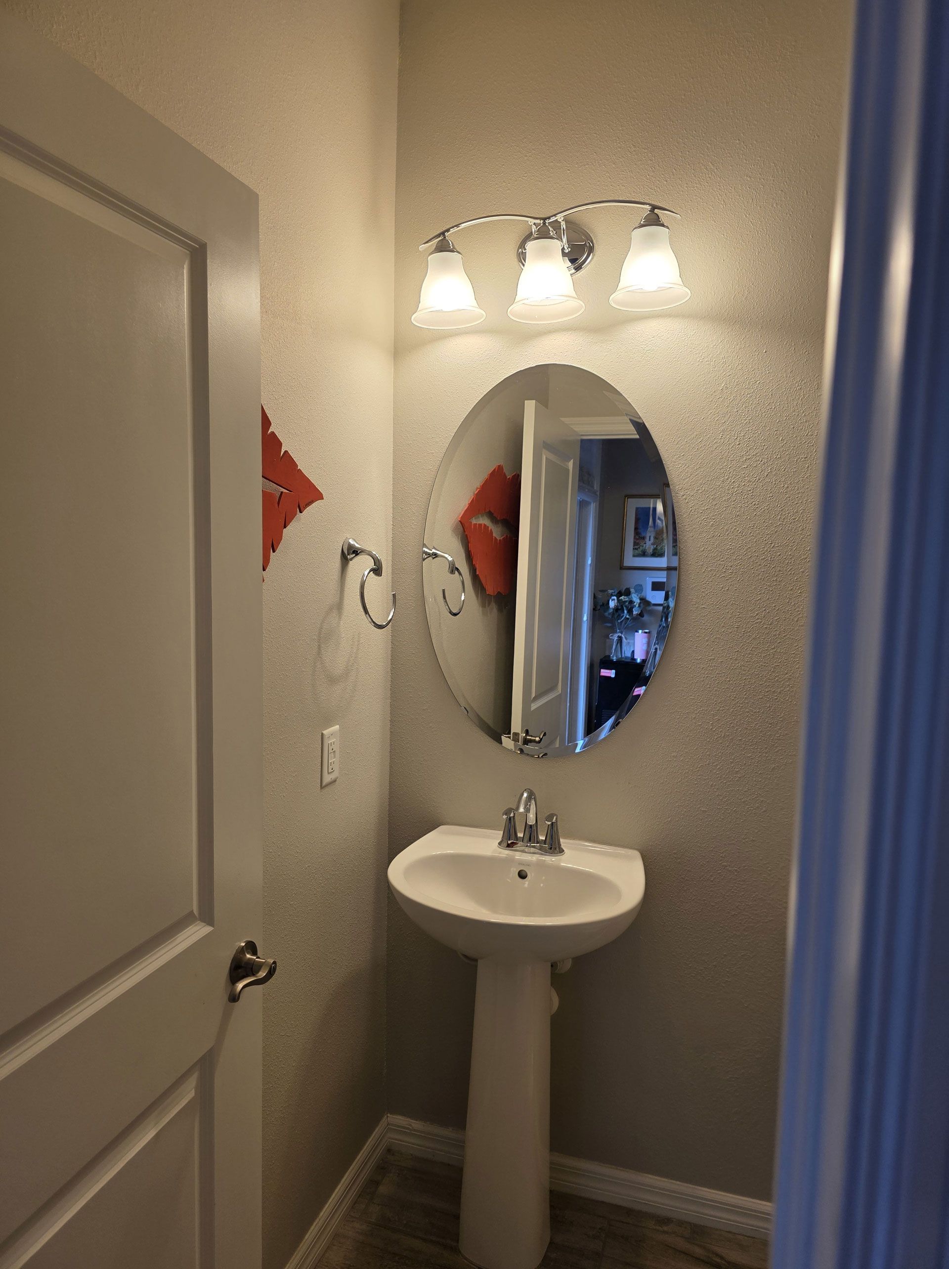 Small bathroom with pedestal sink, oval mirror, and three-light fixture. Gray speckled walls, white door, and red accents.