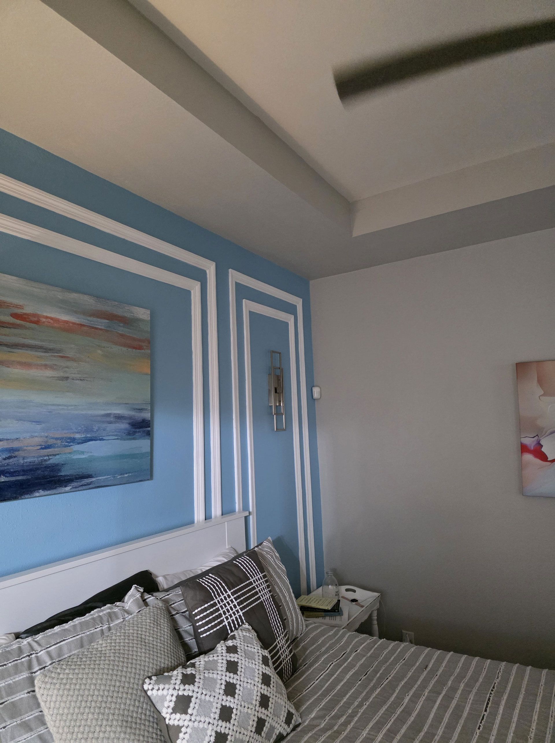 Bedroom with blue wall, white trim, artwork, bed with pillows, and ceiling fan.