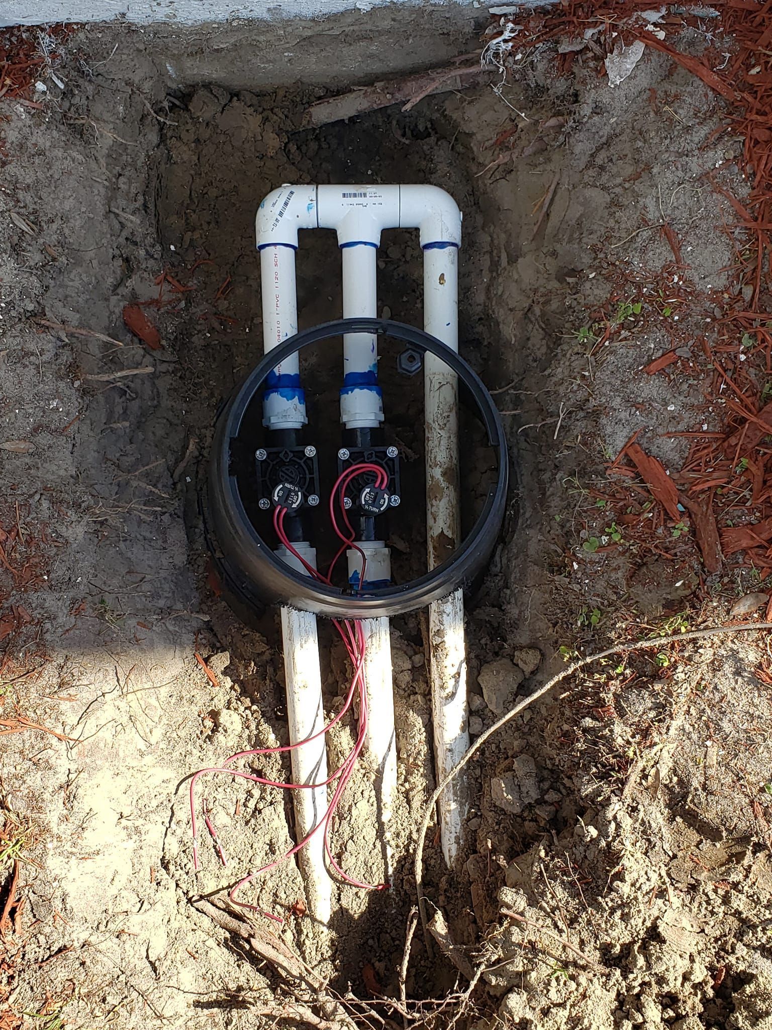 Irrigation system valves in an open black box, with white PVC pipes and wiring, set in a dirt patch.