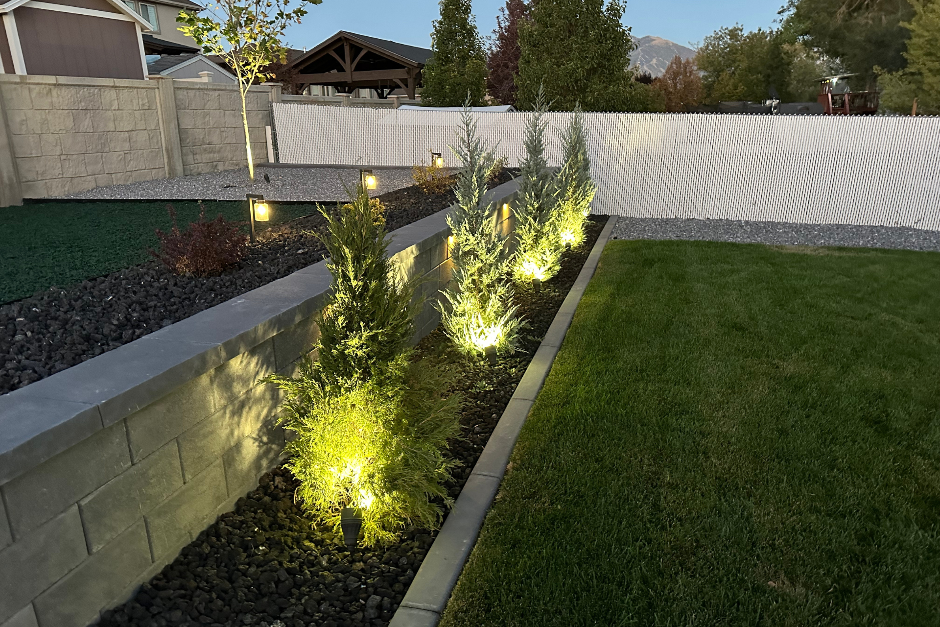A retaining wall with a row of plants are lit up in a garden with a white fence in the background.