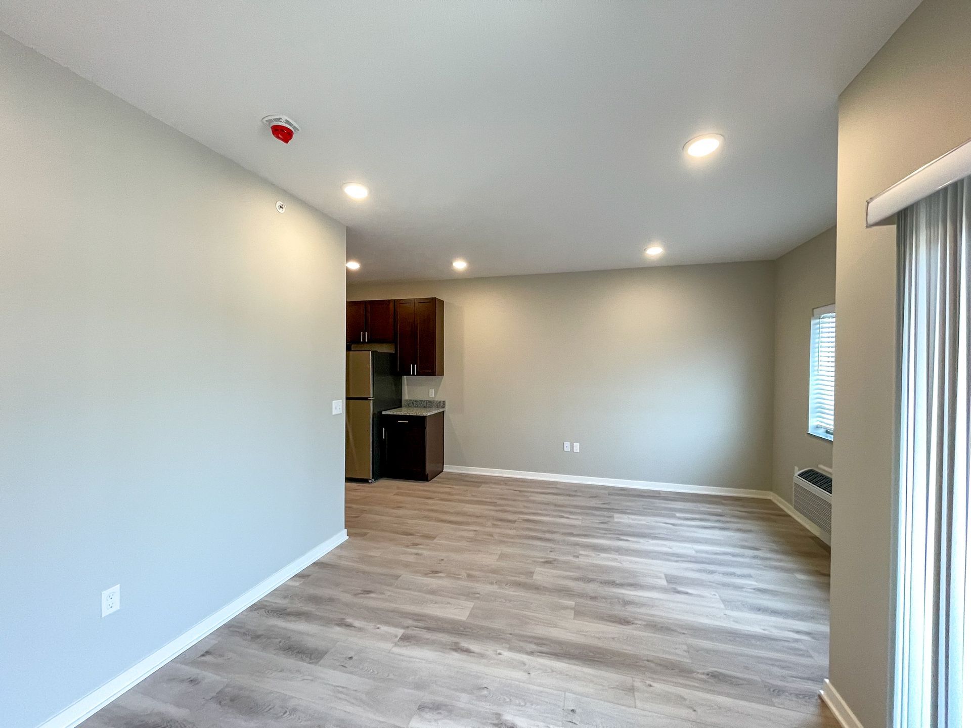 Empty modern living room with gray walls, wood flooring, recessed lights, and a kitchen nook in back.