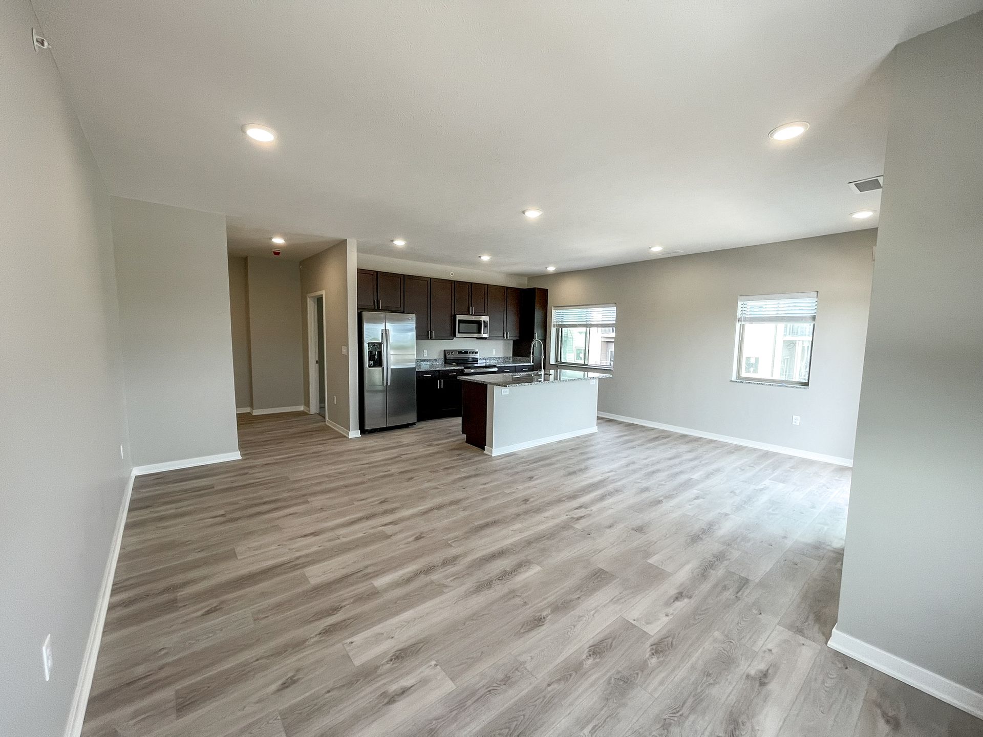Empty open-plan living room with light wood floors and a kitchen island, bright windows, and recessed ceiling lights