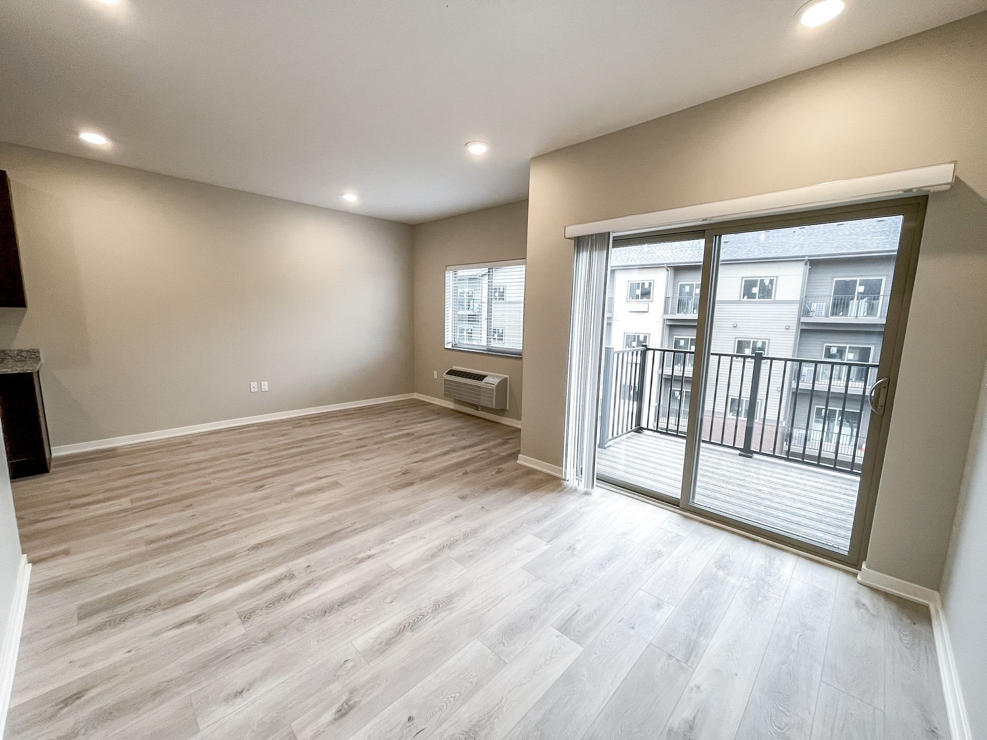 Empty beige living room with light wood floors, recessed lights, a small window, and a sliding glass door to a balcony