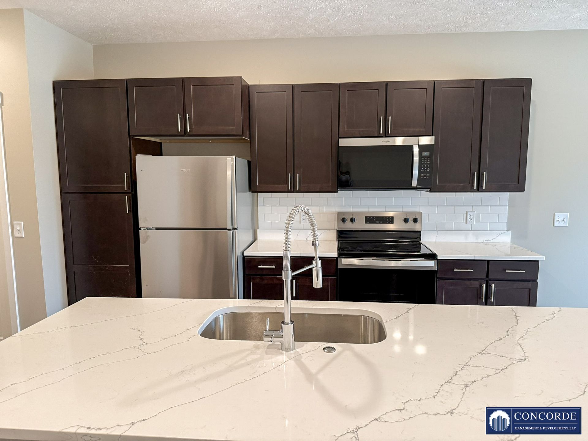 Modern kitchen with dark cabinets, stainless appliances, and a white island countertop with sink.