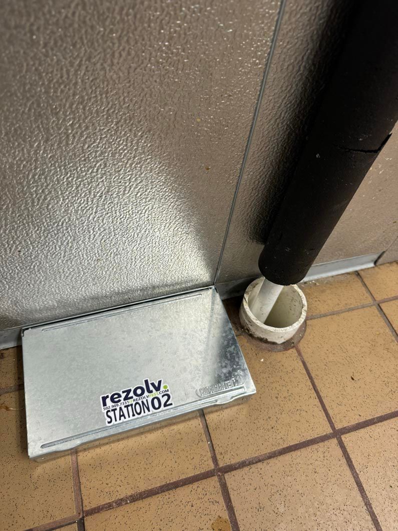 A metal box is sitting on a tiled floor next to a pipe.