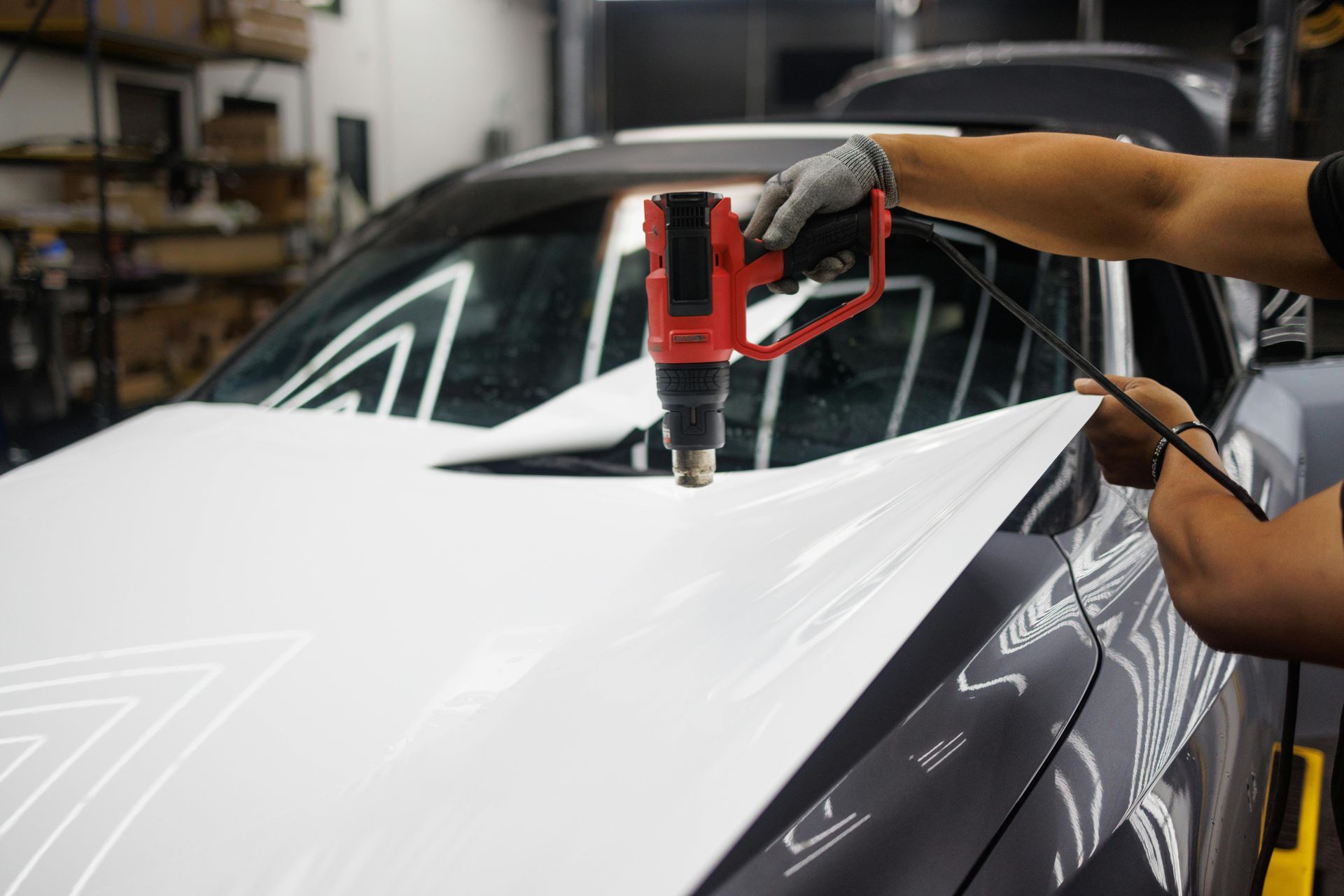 Person applying white vinyl wrap to a car hood with a heat gun.