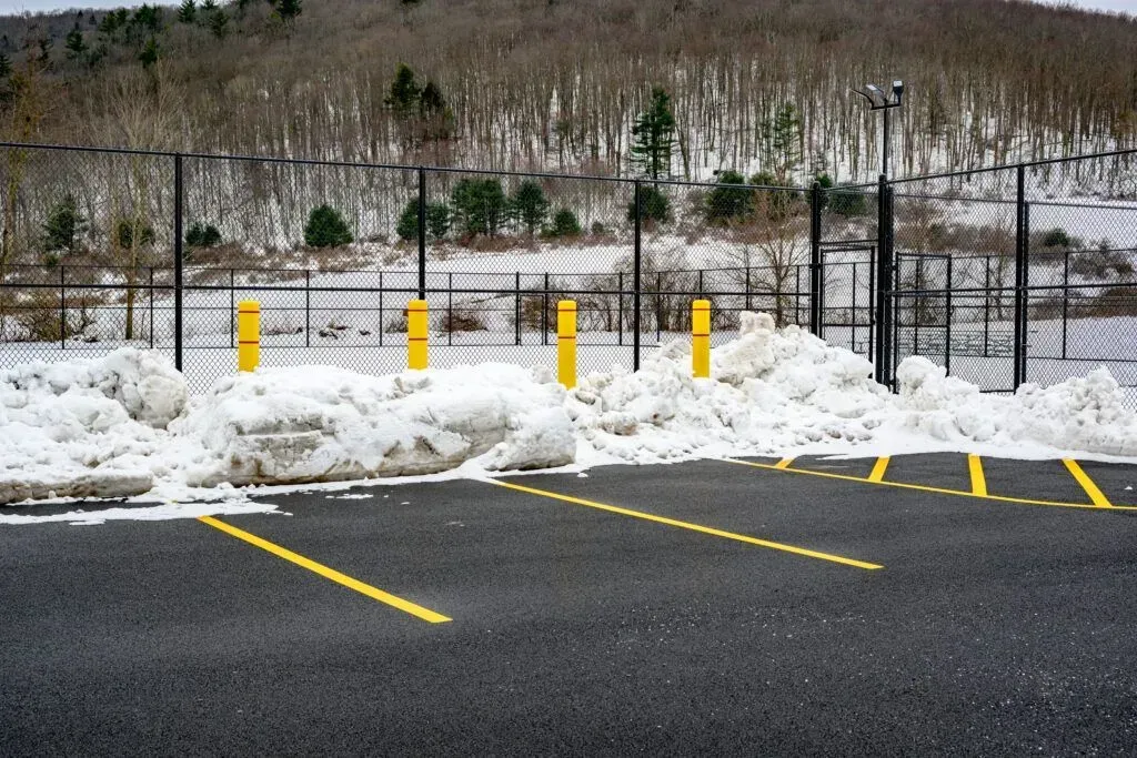 Snowy parking lot with yellow parking space lines and bollards, black fence, and bare trees in the background.