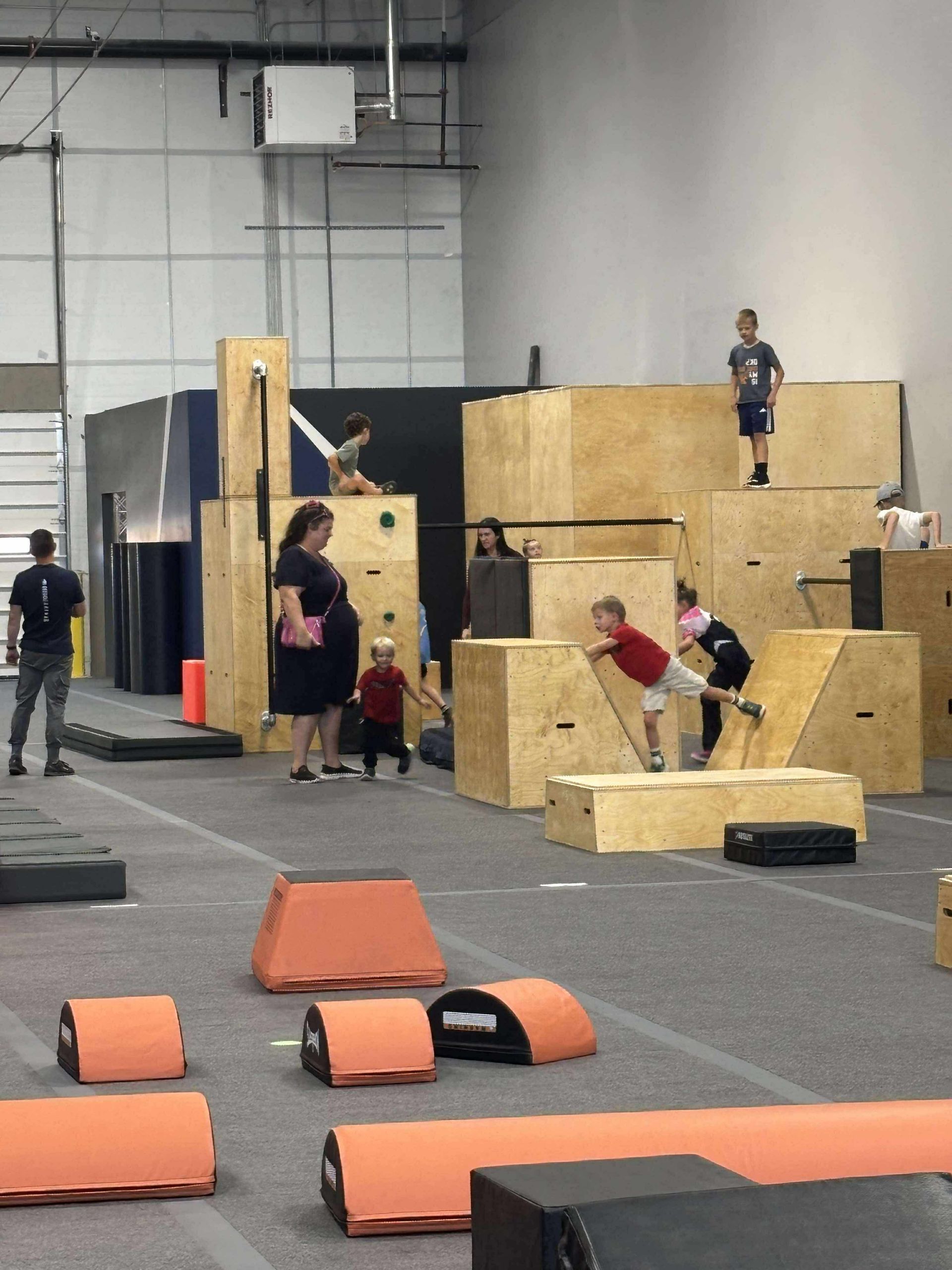 Children playing on parkour obstacles in an indoor gym. Wooden platforms, orange mats.