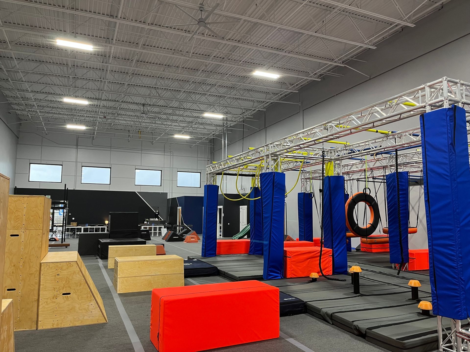 Indoor obstacle course with blue, orange, and wood-colored structures. Ropes, rings, and padding are present.