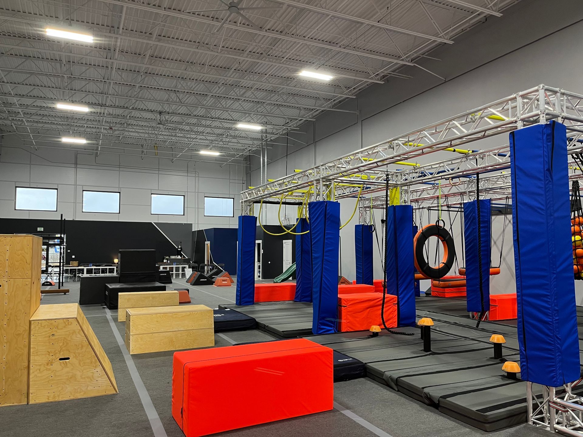 Indoor obstacle course with blue and orange structures, gray mats, and a metal overhead rig.