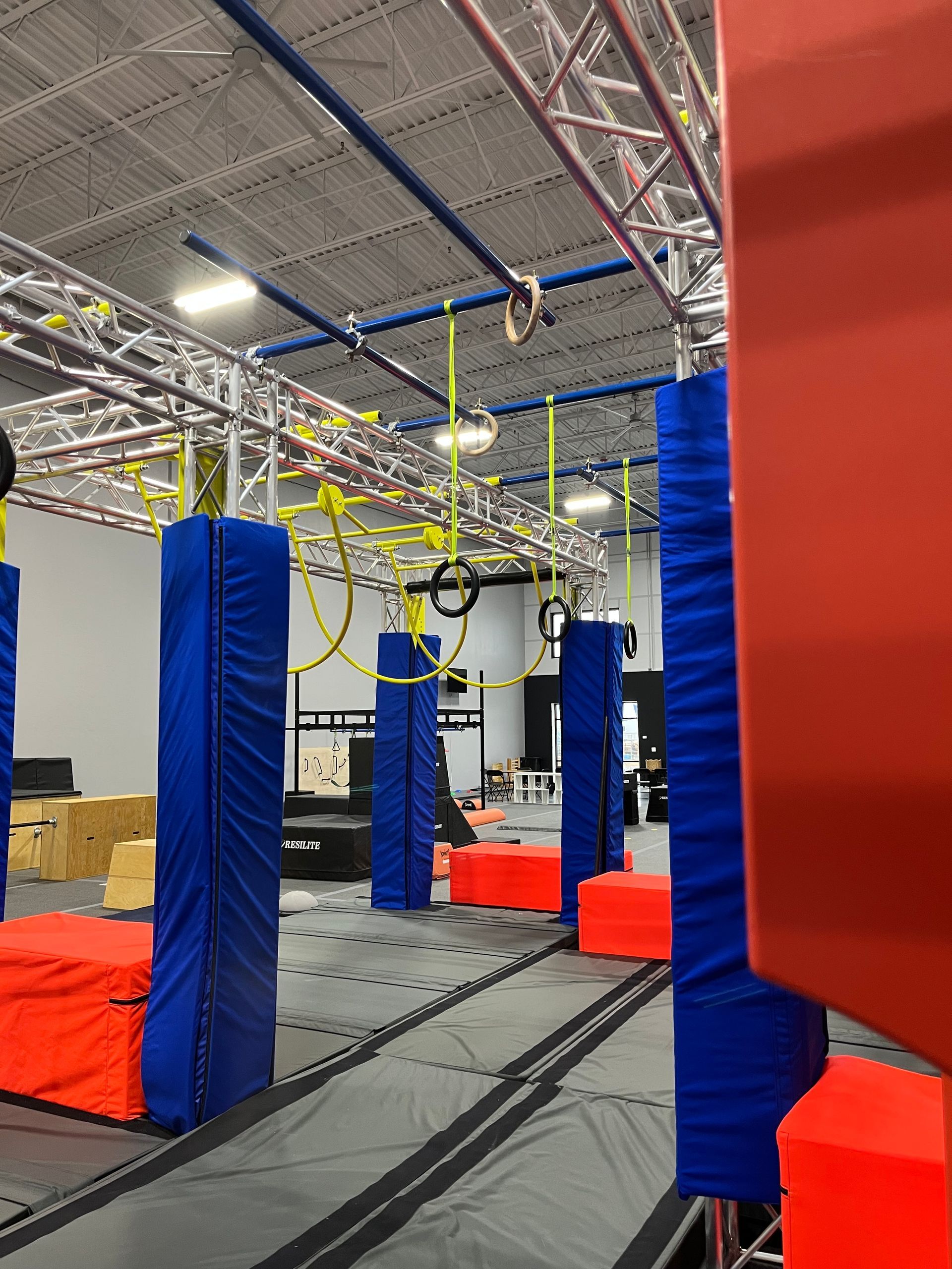 Indoor ninja warrior course with blue and red obstacles, hanging rings, and overhead structures.