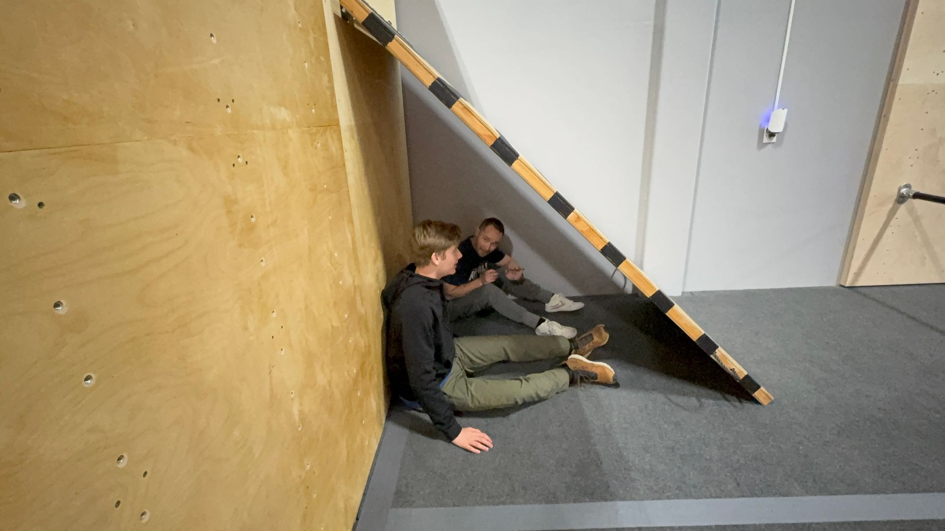 Two people sit on the floor by a wall, under a wooden structure. Indoor setting with neutral colors.