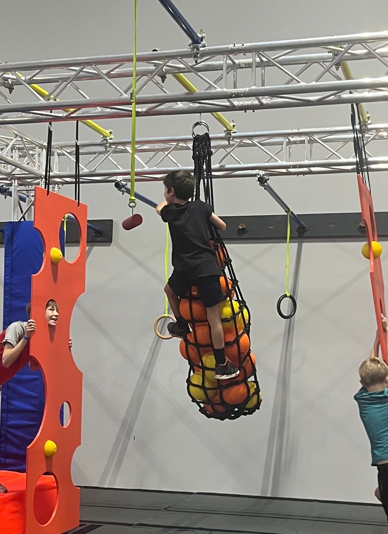 Child climbing a cargo net filled with balls at an indoor obstacle course.
