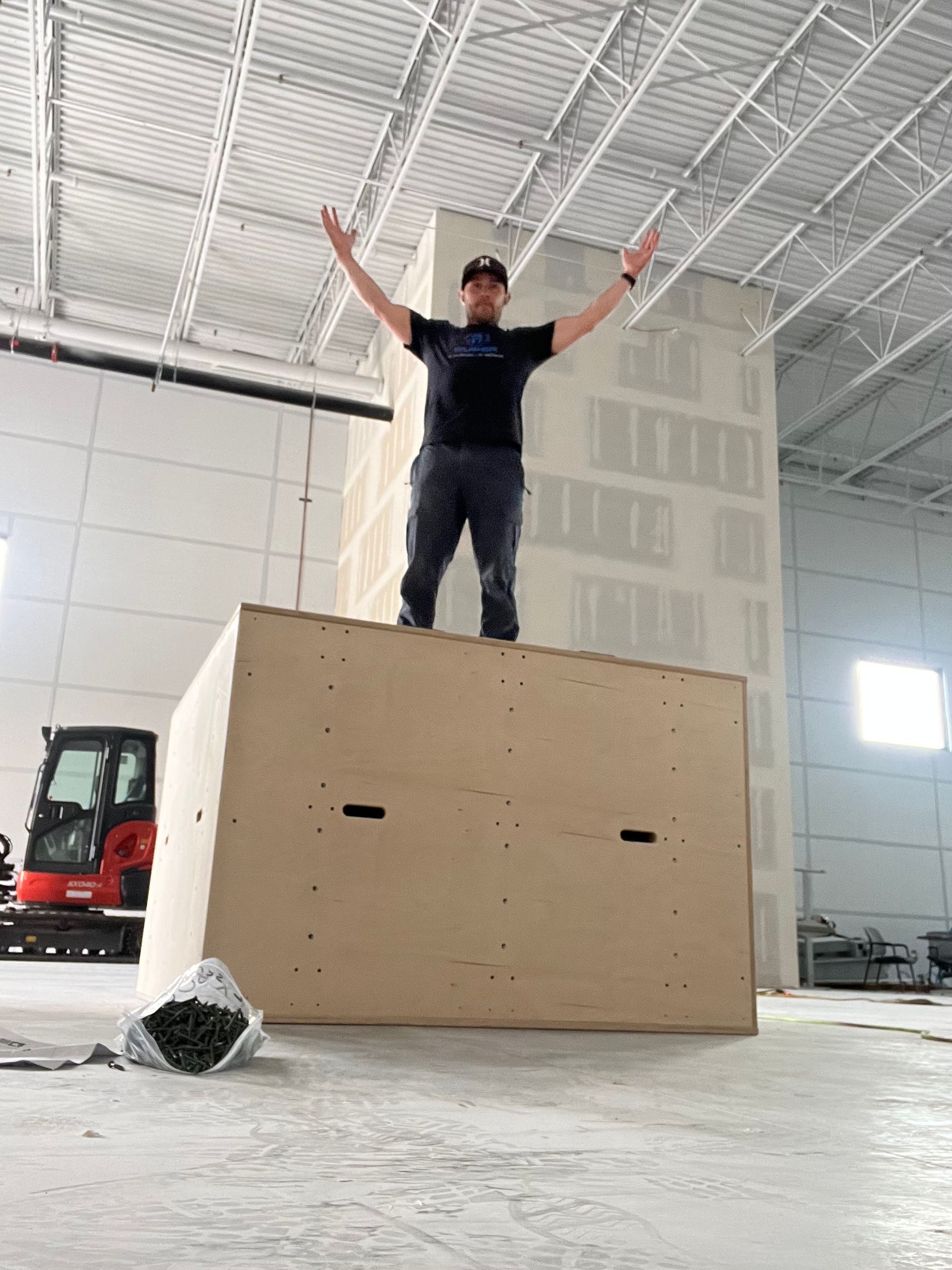 Man stands atop a large tan box with arms raised in a construction setting.