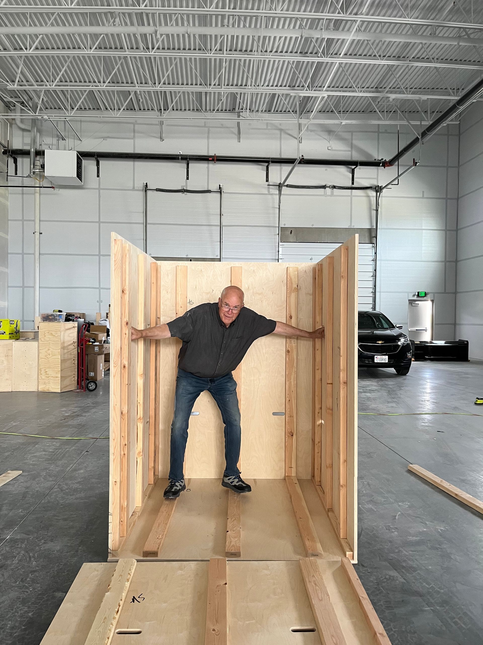 Man standing in wooden structure with arms outstretched in a warehouse setting.