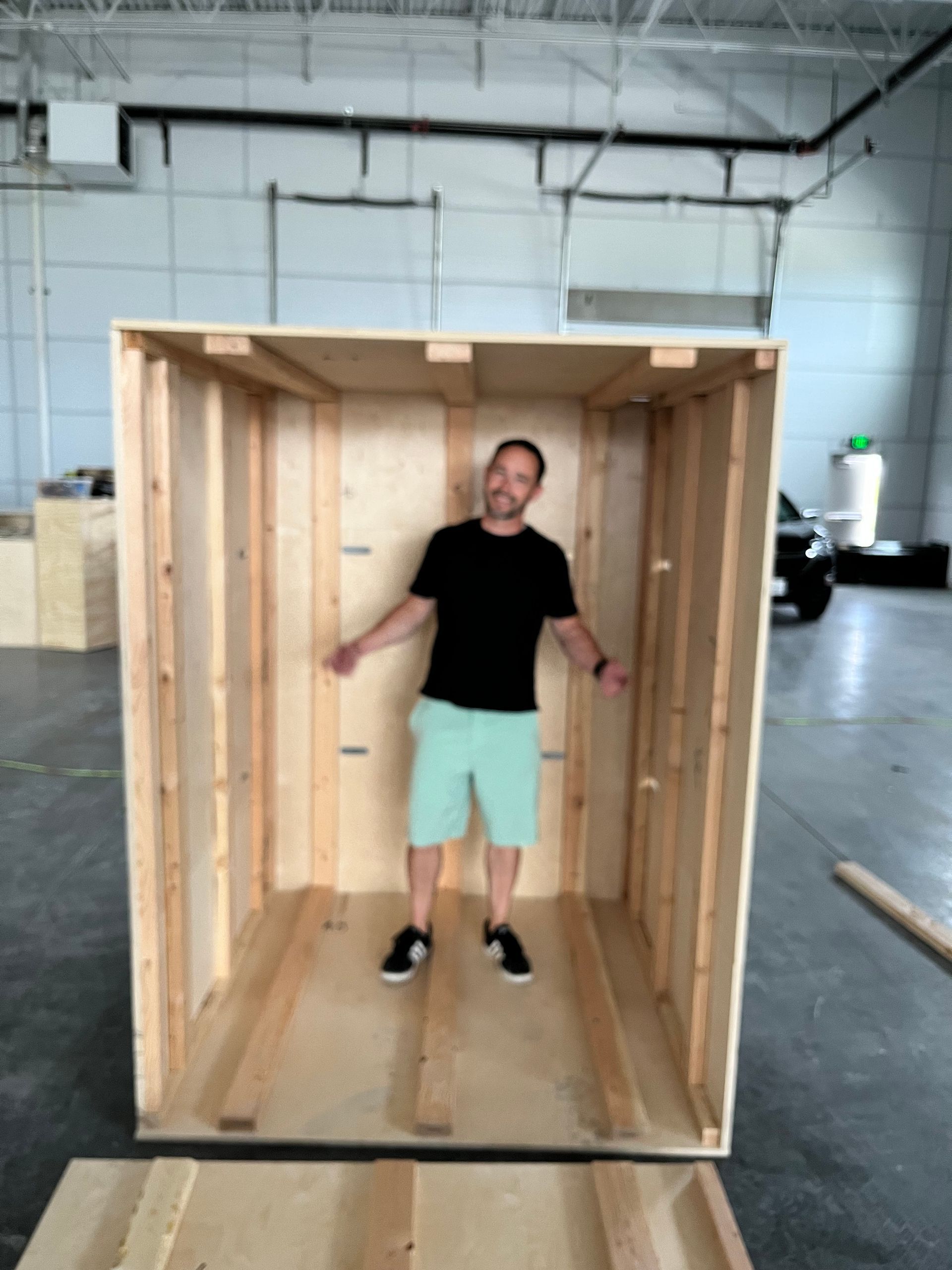 Man standing inside a large, open wooden box in a warehouse. He wears a black shirt, green shorts, and a smile.