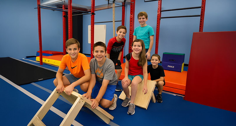 Six children in a gym posing next to wooden obstacle. Blue and red gym setting.