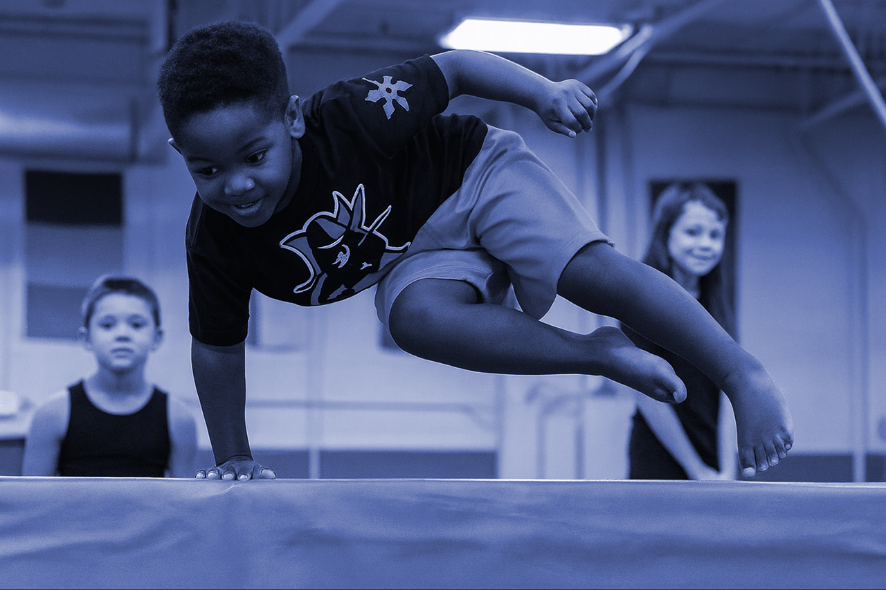 Child doing parkour, balancing on a low bar. Indoor gym setting, other children watching.