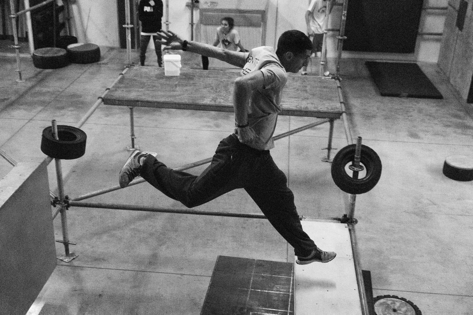 Man leaping over obstacles in an indoor training space, arms outstretched, leg extended. Black and white.