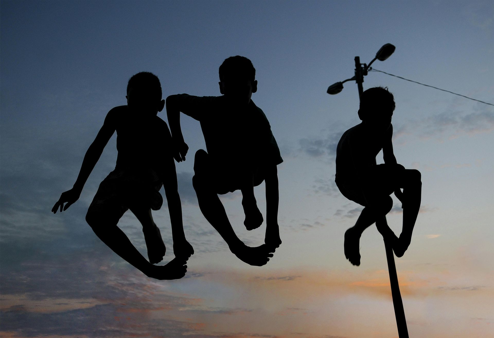 Silhouetted figures of three people; two jumping, one atop a pole, against an evening sky.