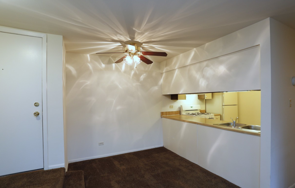 An empty room with a ceiling fan and a kitchen in the background