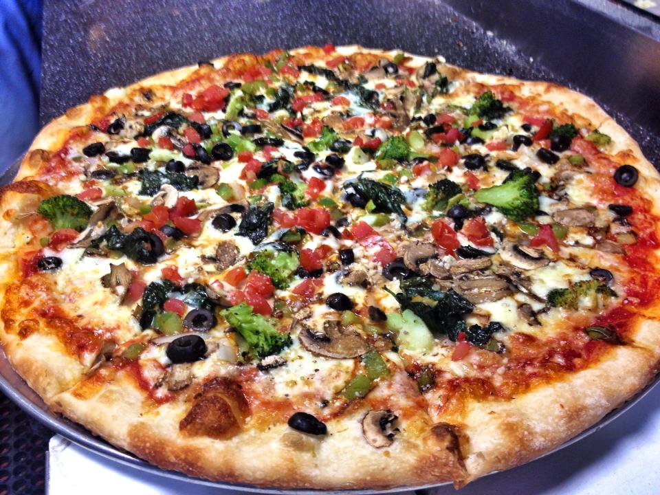 Vegetable pizza on a silver pan; toppings include broccoli, olives, tomatoes, and mushrooms.