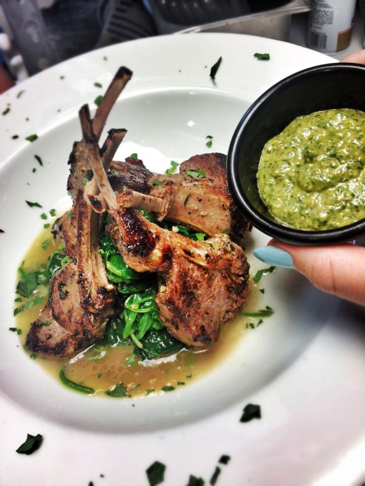 Rack of lamb with green sauce, on spinach bed, served on a white plate, person holding a small bowl of sauce.