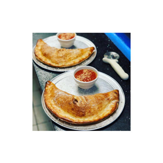 Two golden-brown calzones on metal plates, with marinara sauce and pizza cutter on a countertop.