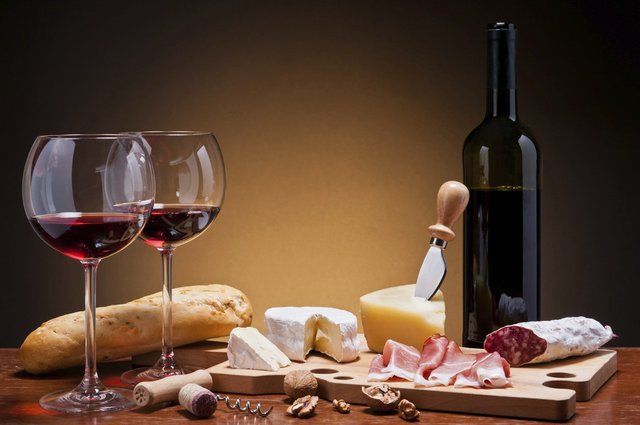 Two glasses of red wine, bottle, cheese, meats, and baguette on a wooden table, with a warm background.