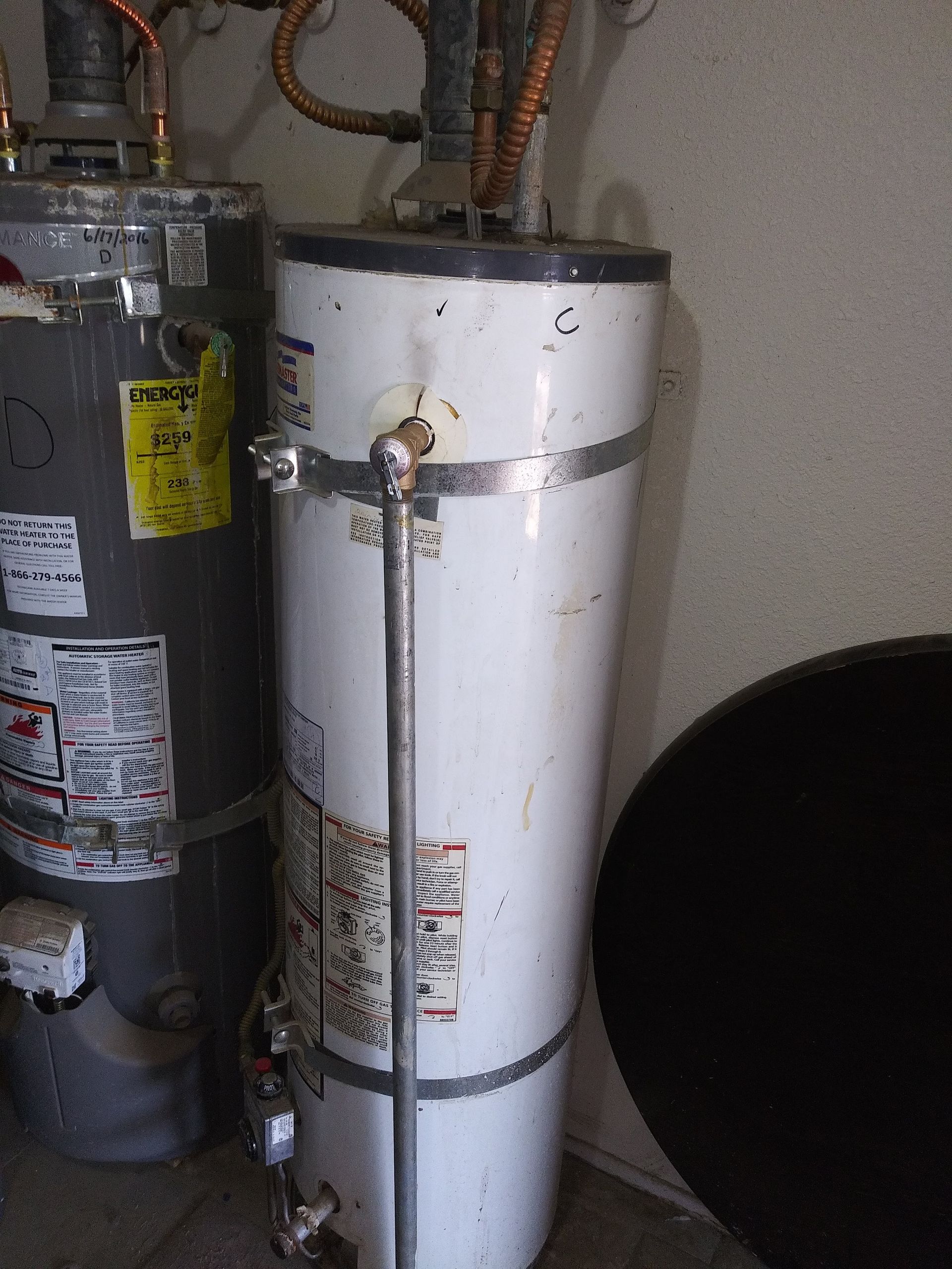 Two water heaters; one white, one gray, in an indoor setting. Pipes and labels visible.