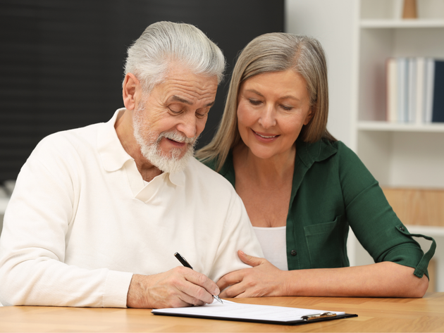 Older person signing a document with a pen, assisted by another person, indoors.