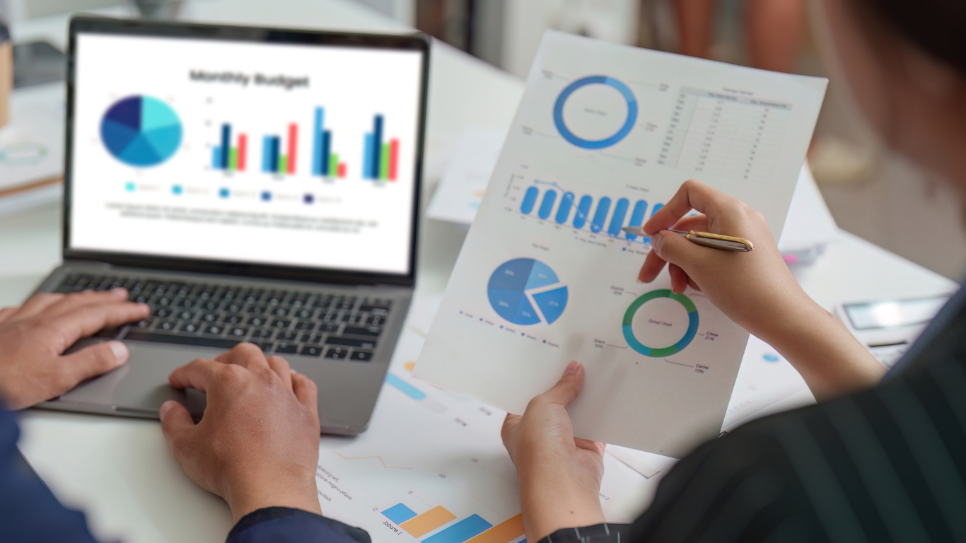 Two people analyzing charts and graphs on a laptop and printed papers, discussing monthly budget.