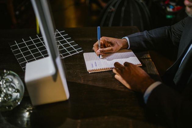 Person writing in a notebook, lit by a lamp, on a dark wooden surface.