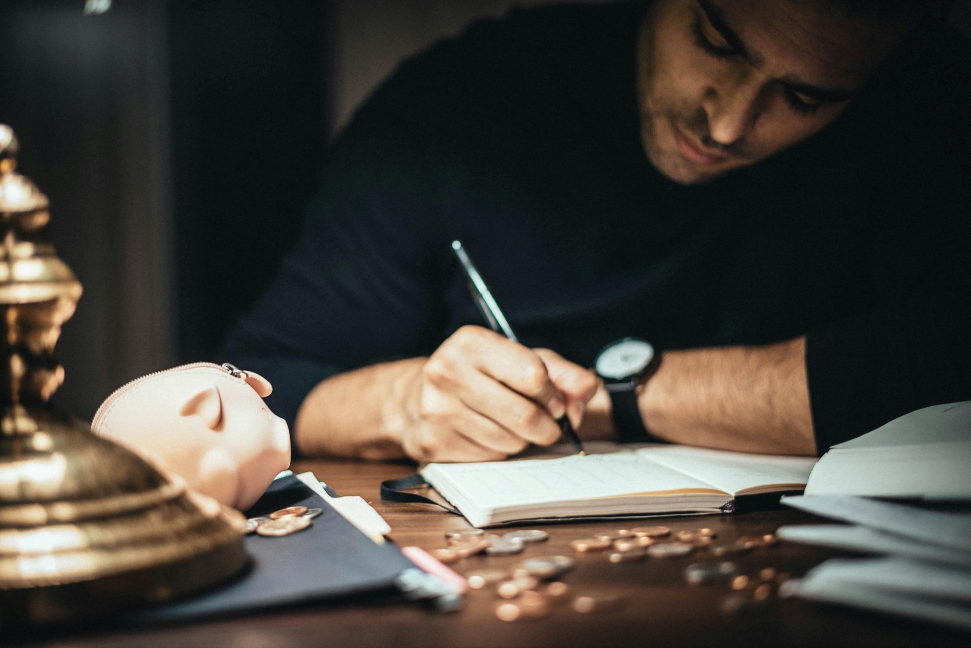 Man writing in a notebook next to a piggy bank and scattered coins on a desk in a dimly lit room.