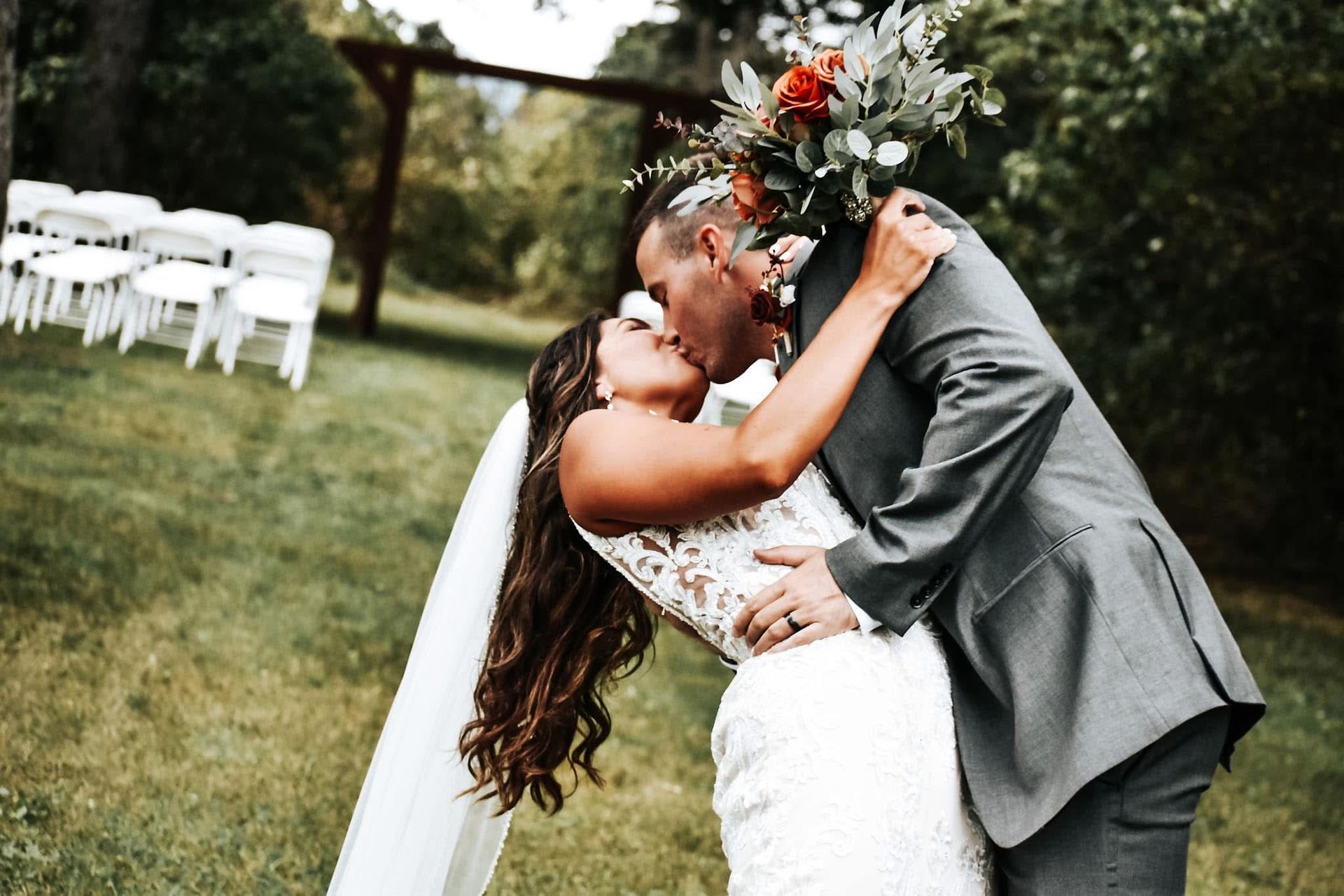 A bride and groom are kissing in the grass at their wedding.