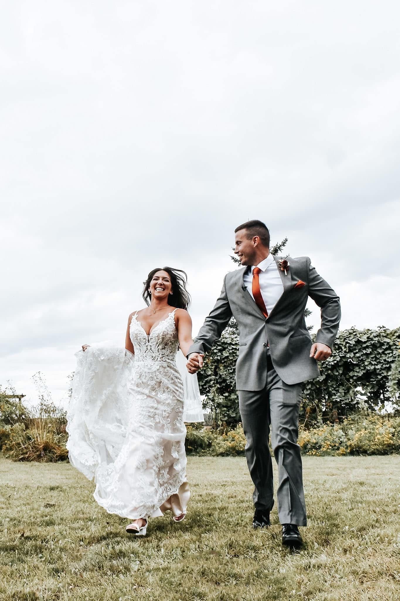 A bride and groom are walking through a grassy field holding hands.