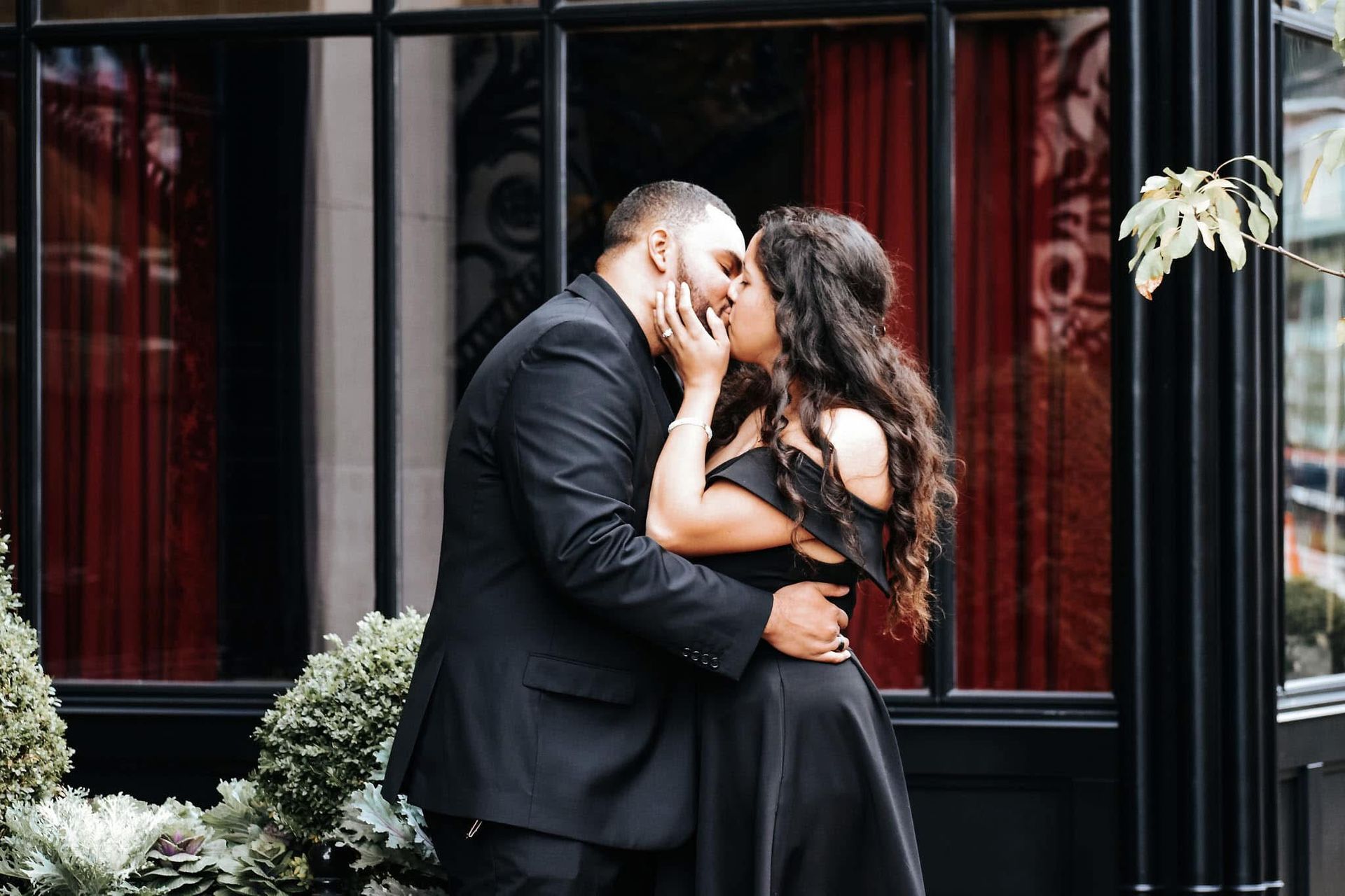 A man in a suit and a woman in a black dress are kissing in front of a window.