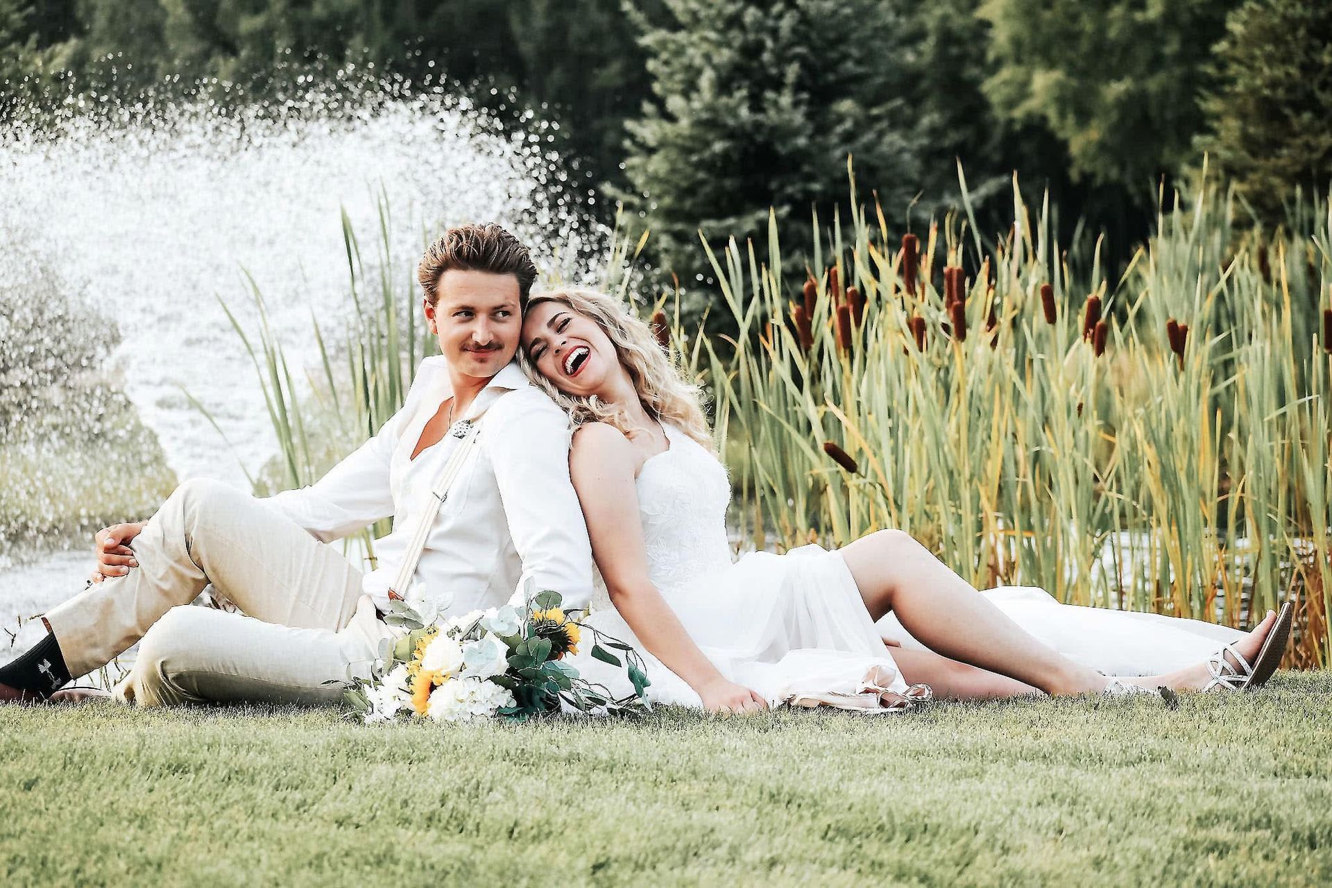 A bride and groom are sitting on the grass in front of a fountain.