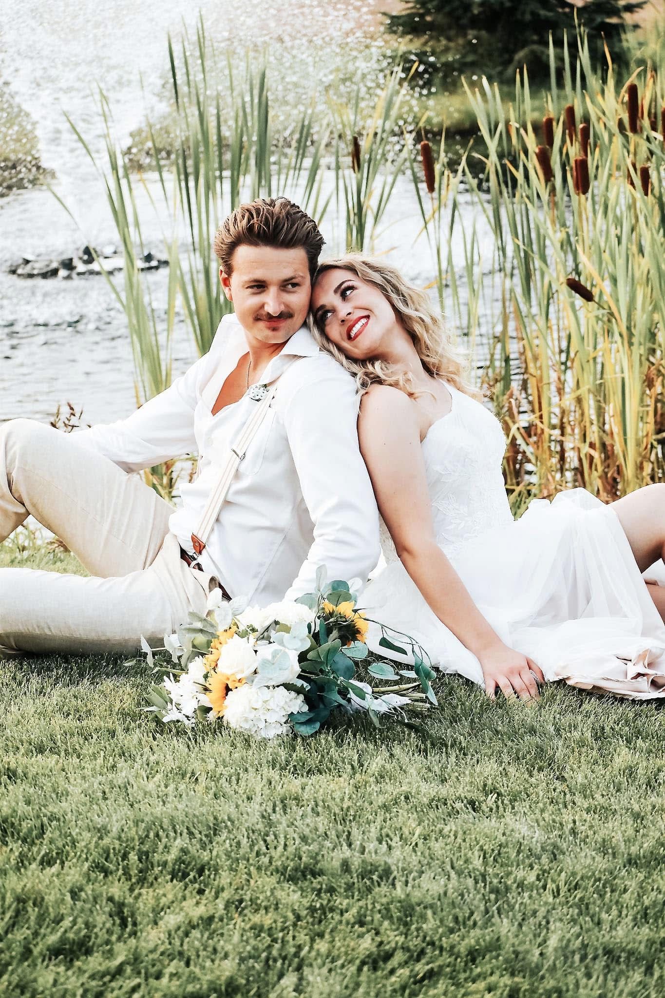 A bride and groom are sitting in the grass near a pond.