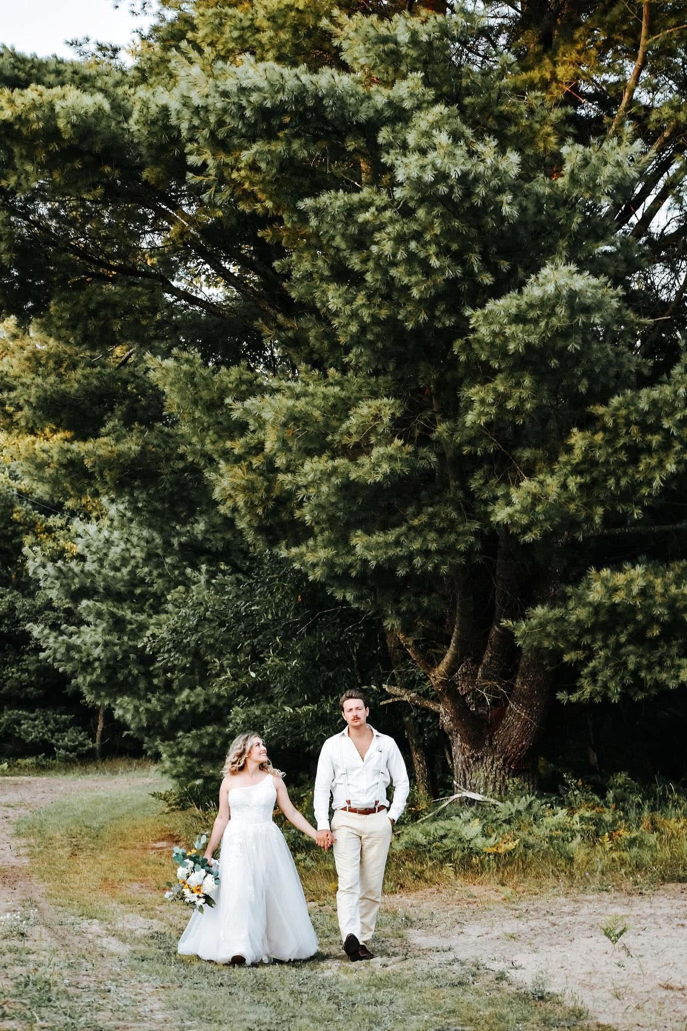 A bride and groom are walking down a path in the woods holding hands.