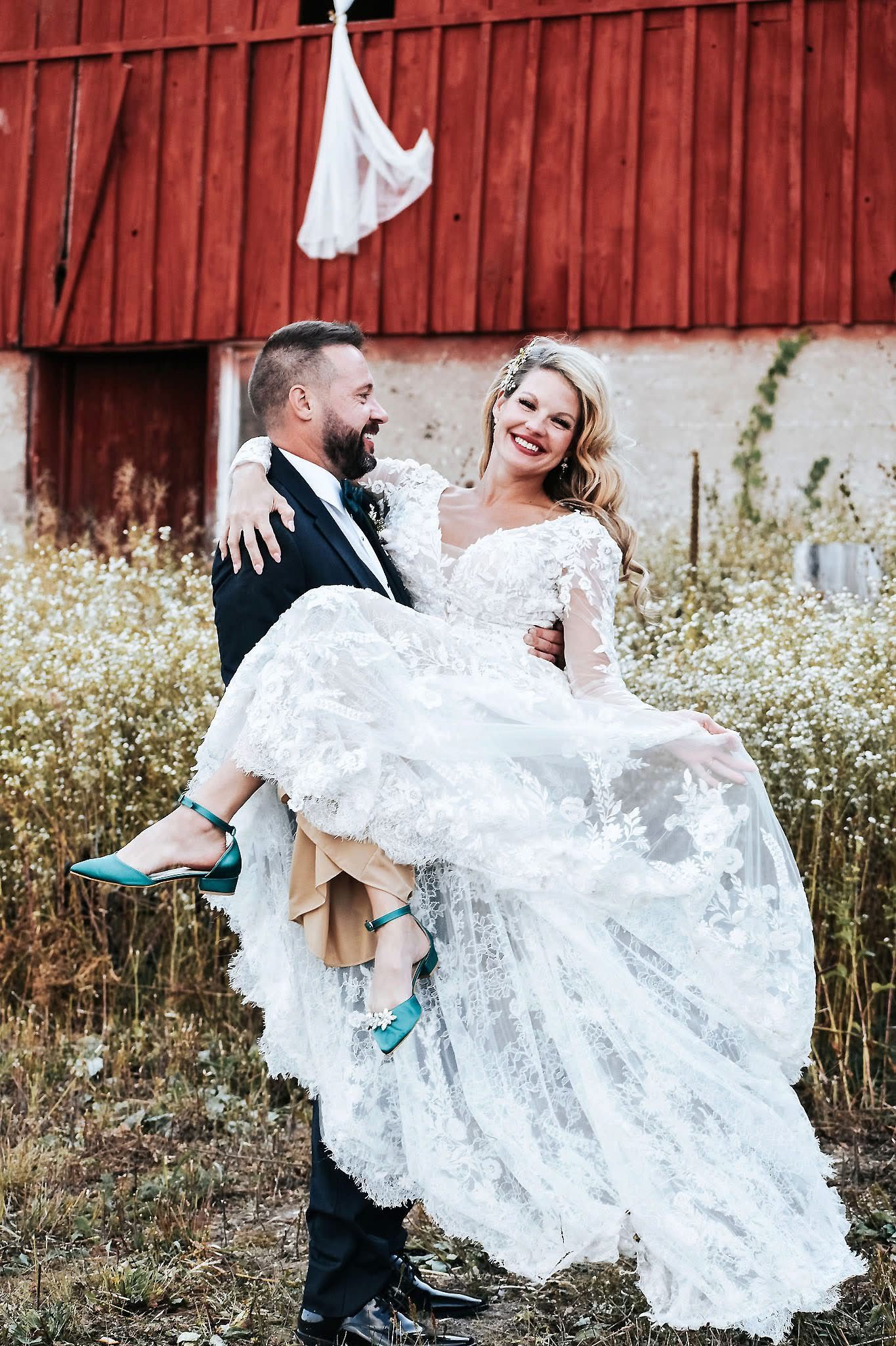 A man is holding a bride in his arms in front of a red barn.