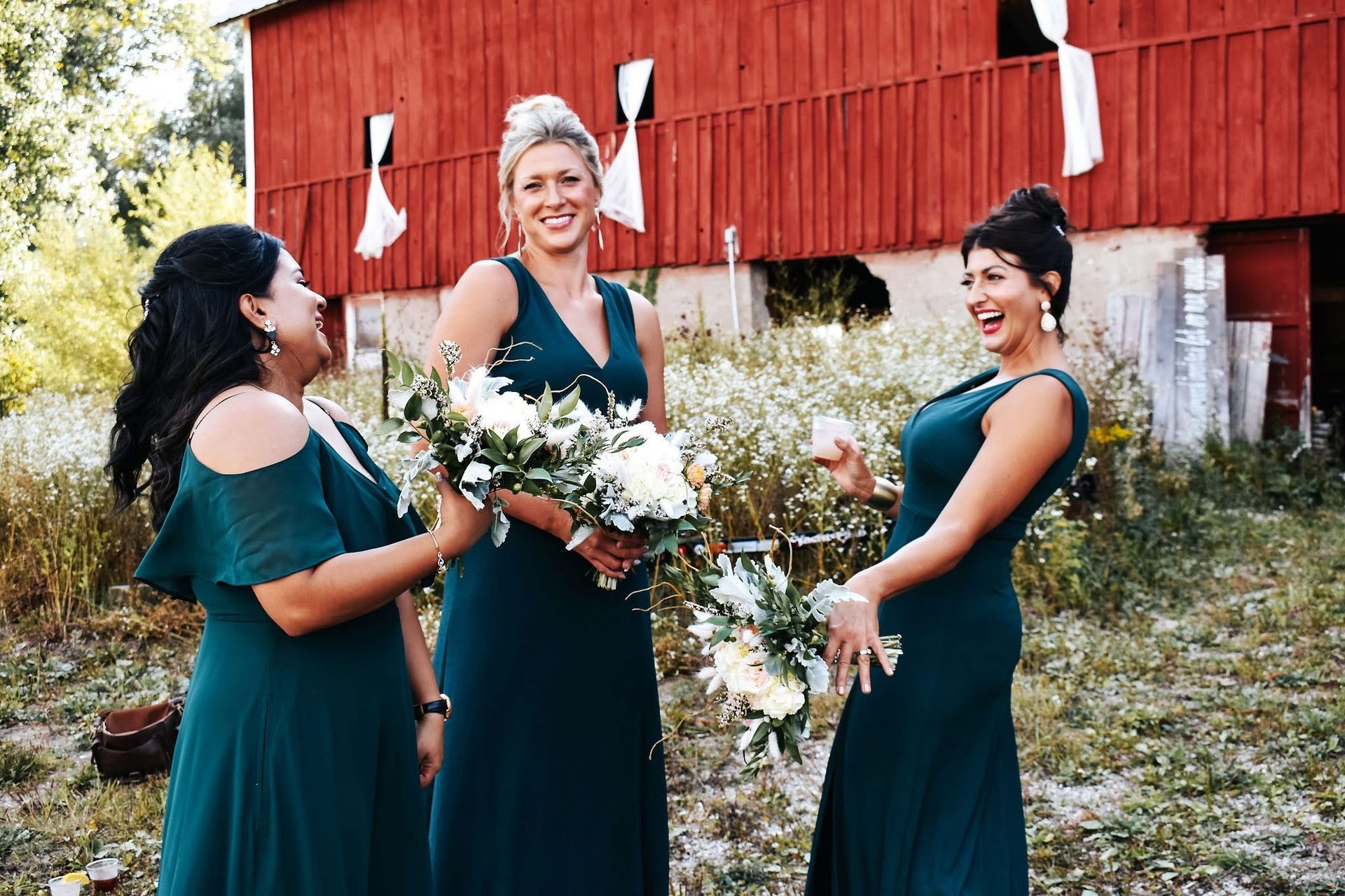Three bridesmaids in green dresses are standing in front of a red barn.