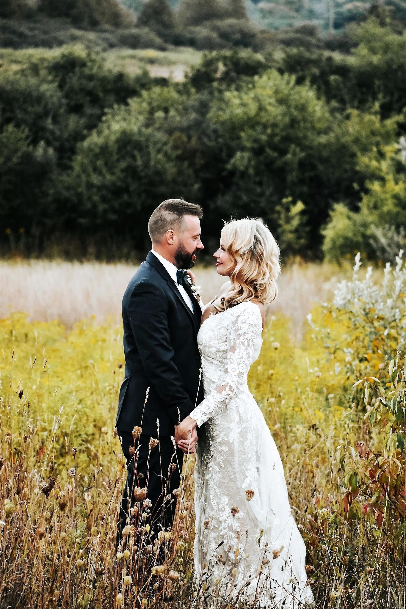 A bride and groom are standing in a field holding hands and looking at each other.