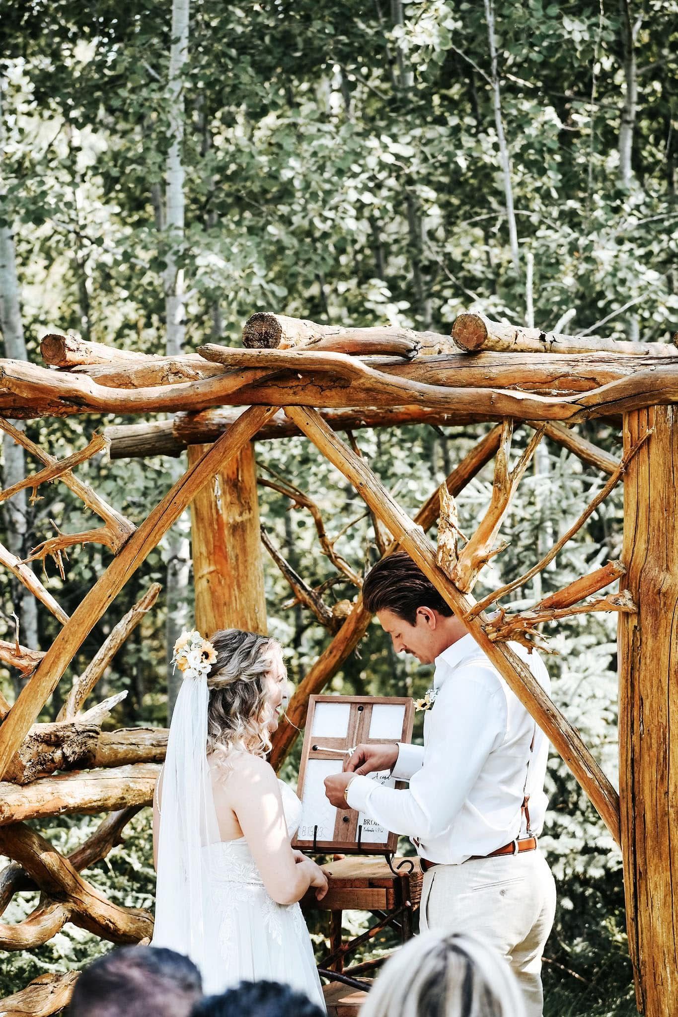 A bride and groom are getting married under a wooden archway in the woods.
