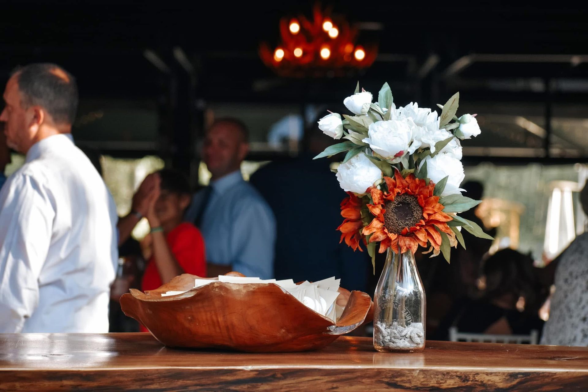 A vase of flowers sits on a wooden table in front of a crowd of people.
